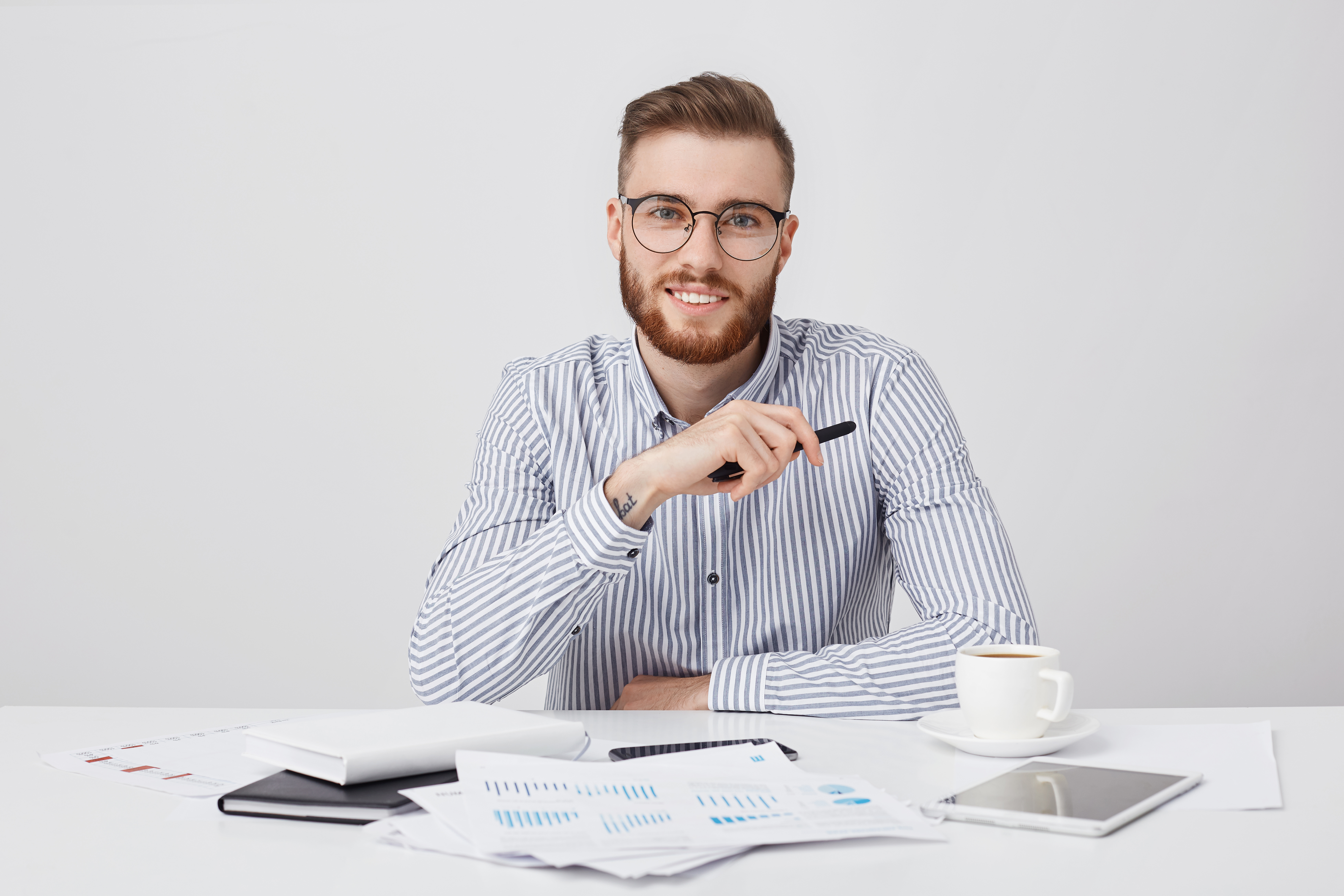 Professional male worker with thick beard and trendy hairstyle, wears round glasses and formal shirt, sits at work plae, drinks coffee, works with documents. Hipster young guy being busy working